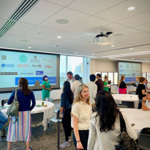 A group of people are networking in a bright, modern conference room with round tables and large windows. Two projection screens display a slide that thanks various partner organizations, and the room has a collaborative, casual atmosphere.